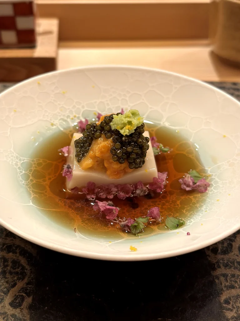 Sesame tofu topped with sea urchin and caviar in a light broth, garnished with edible flowers and citrus zest in an elegant Japanese omakase presentation.