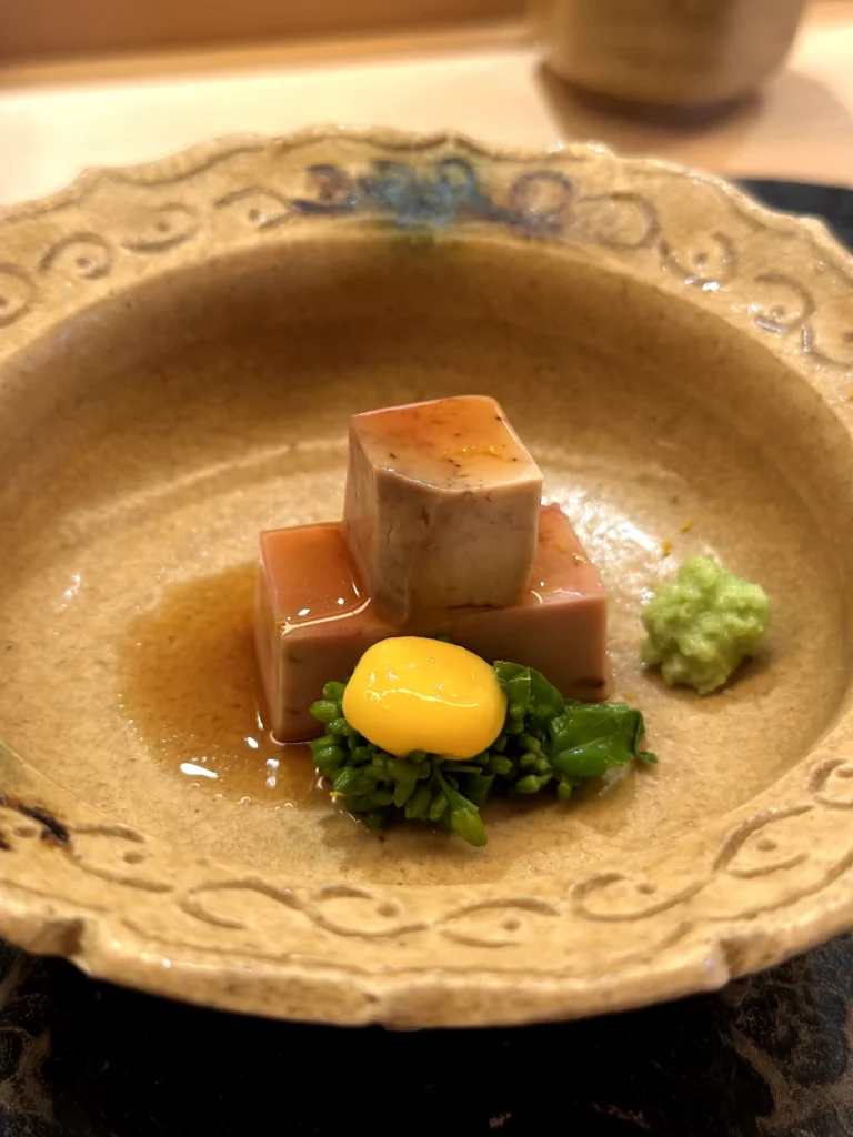 Monkfish liver served in a delicate soy-based sauce with seasonal greens, mustard garnish, and Japanese egg yolk in a handcrafted ceramic bowl.