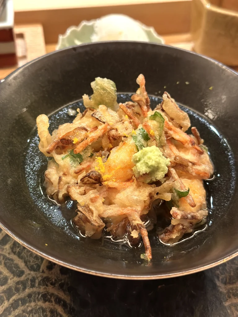 Tendon-style tempura rice bowl with mixed seafood and vegetables in a savory broth, garnished with wasabi and citrus zest.