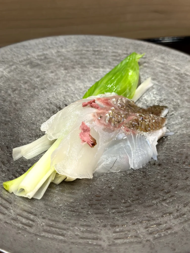 Thinly sliced white fish sashimi garnished with spring onions and edible blossoms on a textured ceramic plate