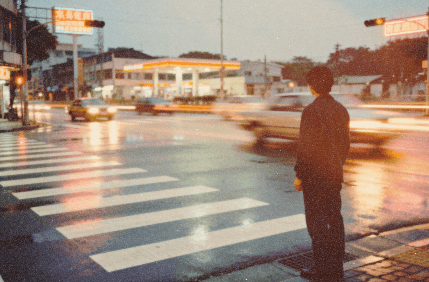 A person stands at a crosswalk on a rainy evening, watching cars pass by with headlights creating light trails. The street reflects the glow from nearby buildings and traffic lights, adding a soft, nostalgic atmosphere.
