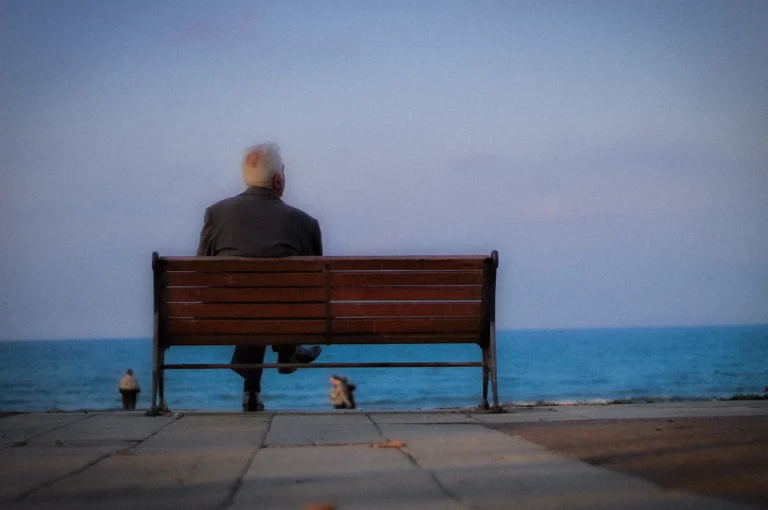 Elderly man sitting alone on a wooden bench overlooking the sea during a peaceful evening, capturing a quiet moment of reflection by the waterfront.