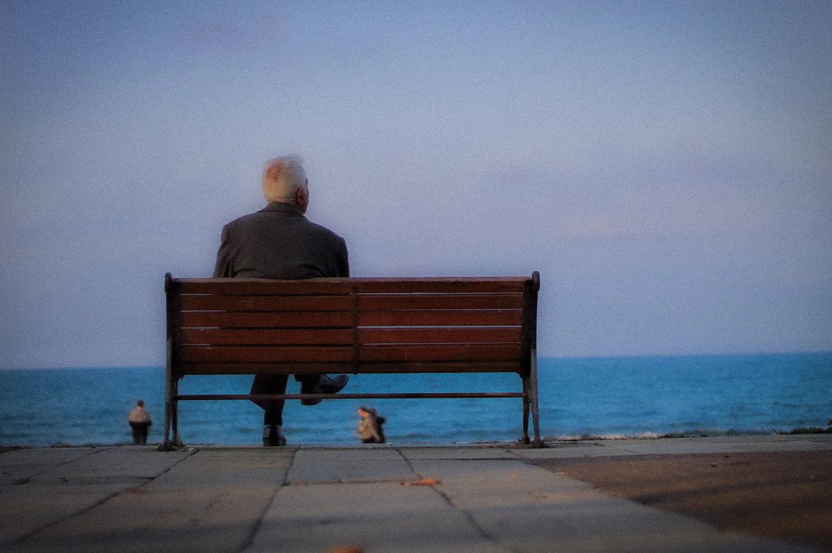 Elderly man sitting alone on a wooden bench overlooking the sea during a peaceful evening, capturing a quiet moment of reflection by the waterfront.