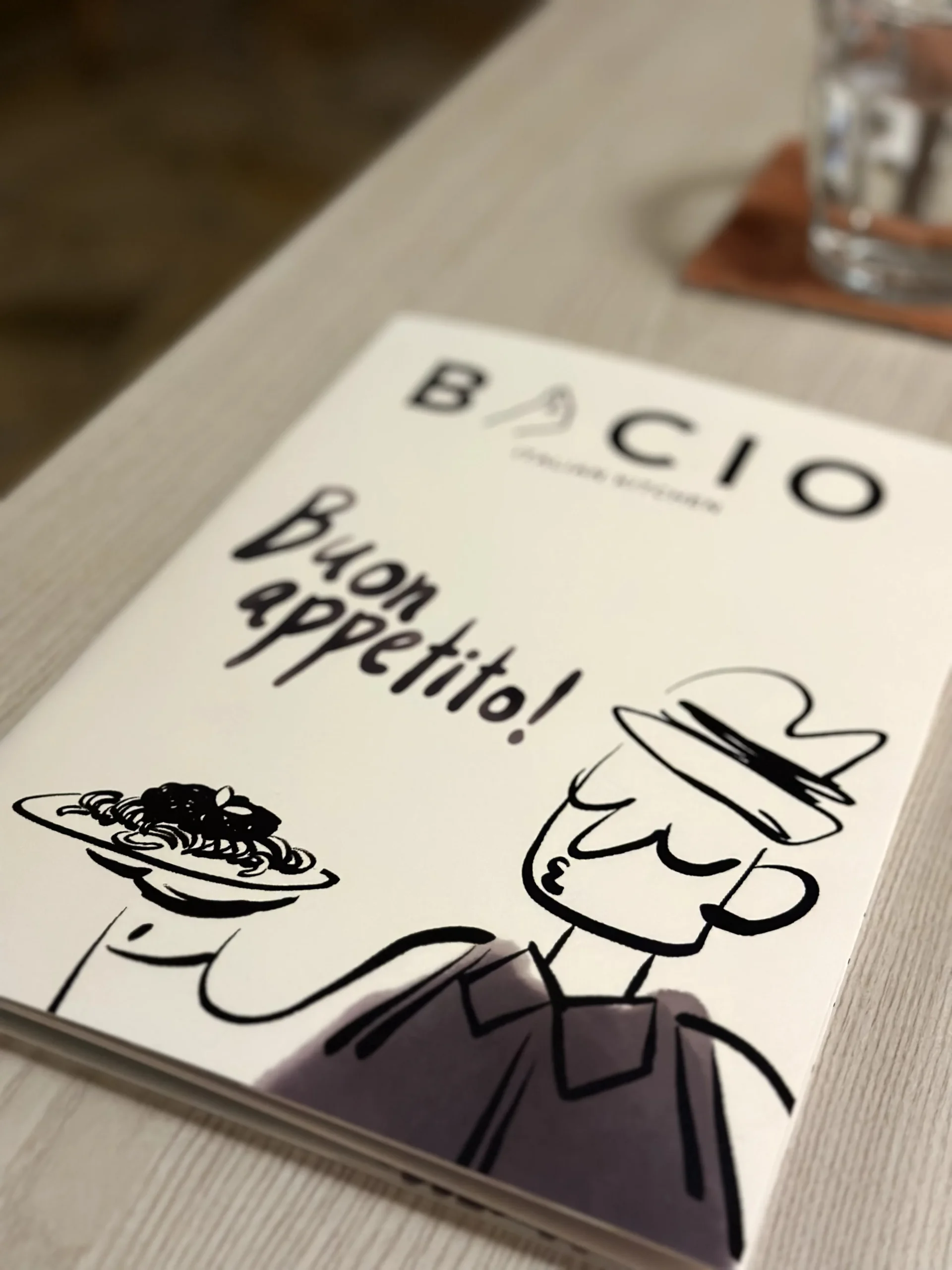Bacio Italian Restaurant menu cover featuring a minimalist chef illustration and “Bon Appetito!” greeting on a modern dining table.